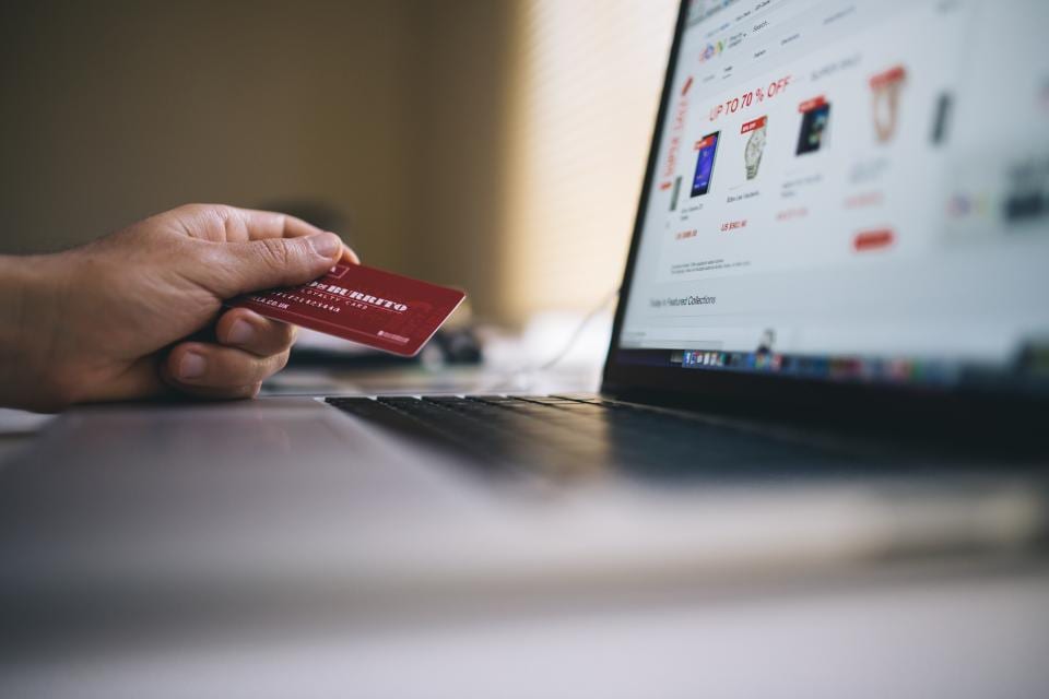 A hand holding a bank card in front of a laptop showing an online shop, a shopper about to buy at the advertised price